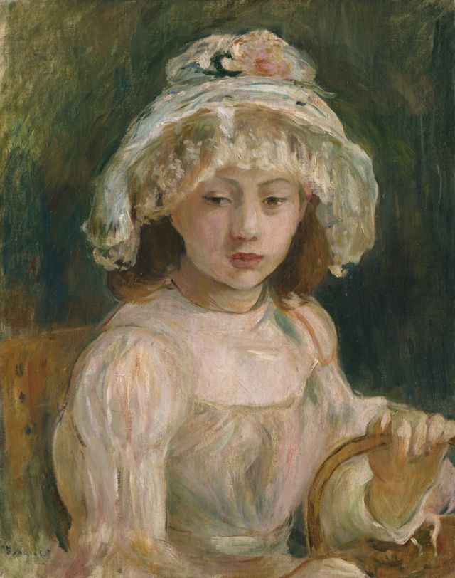Portrait of a young girl in a white ruffled hat and pale pink dress, seated in a wicker chair against a dark green background.